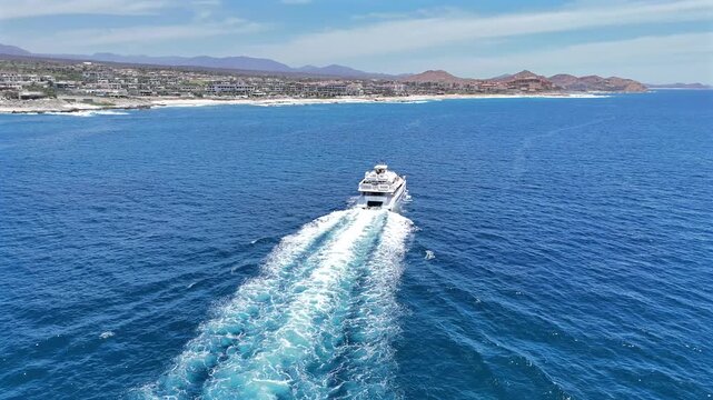 Drone orbits a catamaran yacht moving through the ocean just off the Cabo del Sol cape, with the eastern Cabo San Lucas coastline stretching along the background in bright midday sun.