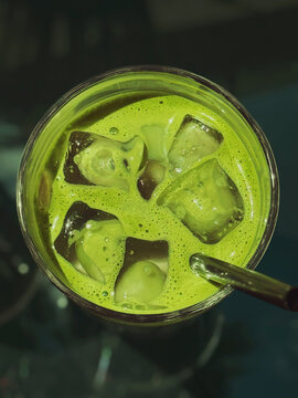 Close-up top view of vivid green iced matcha tea in clear glass with large melting ice cubes and drinking straw. Refreshing cold beverage on dark cafe table