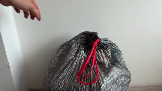 Woman's hand tightly closing a gray plastic garbage bag with red drawstring handles for proper waste disposal, getting ready for collection and recycling at home
