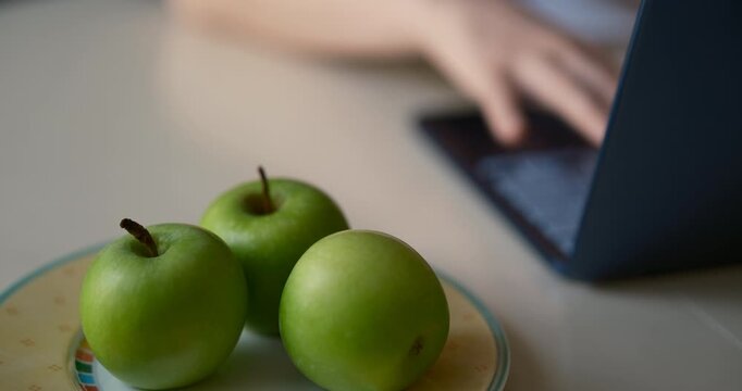 Cinematic close-up of three fresh green apples on a plate beside a laptop while a person works in the background. wellness, remote work, productivity, and balanced lifestyle.