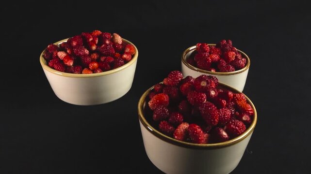Three white bowls filled with ripe red forest wild strawberries rotating on a black background. These freshly picked organic berries showcase a healthy, natural, and delicious dessert ingredient