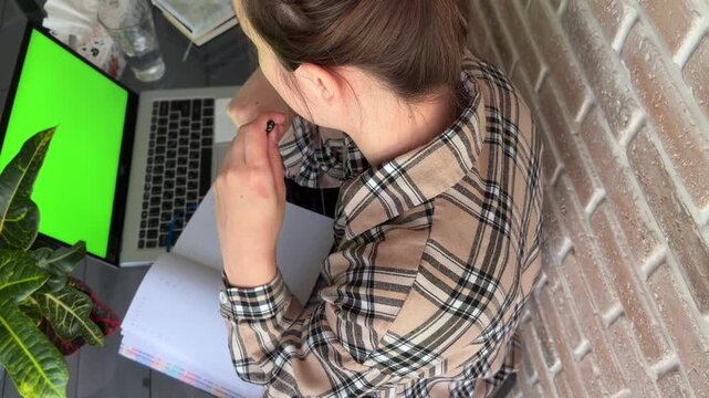 Female student studying online from home using a laptop with a green screen, writing notes in a notebook, taking a break, and rubbing her eyes, showcasing distance education