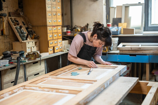 Interested concentrated woman crafter refurbishing vintage wooden door in restoration workshop, craftswoman sanding spackled details and surface using sandpaper, creative working process in carpentry.
