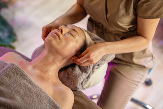 Tranquil woman on spa table receiving skin care treatment and healing massage therapy procedure in salon, masseuse in beauty spa clinic massaging female patient ears using relaxation technique.