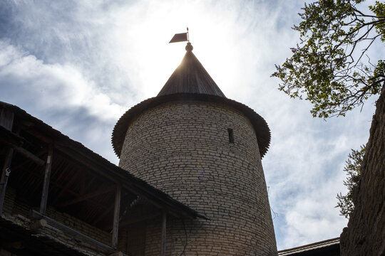 Historic medieval stone fortress watchtower with a wooden conical roof and weather vane, backlit by the bright sun in a blue cloudy sky.
