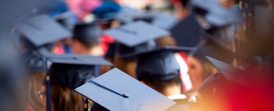 Graduates wear caps in a sea of dark tones. Focus highlights one cap with a tassel in crisp detail. Blurry crowd behind suggests celebration and pride. Warm light glows from right