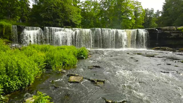 Stunning low altitude aerial view flying over a river towards the scenic Keila Joa waterfall, a beautiful natural landmark surrounded by a lush green forest on a bright summer day