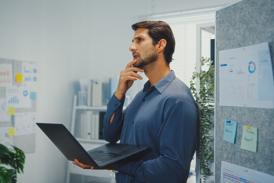Thoughtful young Latin businessman stand with laptop analyzing data above paperwork stack. Hybrid work audit, digital transformation strategy, business corporate finance, executive decision.