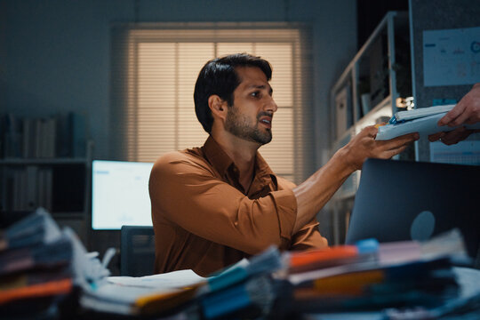 Overworked Latin businessman receiving extra paperwork from boss at desk in dark night office. Working late overload, deadline burnout, heavy overtime stress, corporate business problem concept.