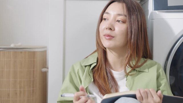 Beautiful, happy Asian woman sitting on floor, leaning against washing machine, looking out the window, thinking and writing diary with smile in sunlight in cozy laundry room at modern home