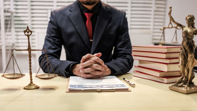 Lawyer sitting at a desk with legal books, scales of justice and a statue, with hands folded and paperwork in front of him