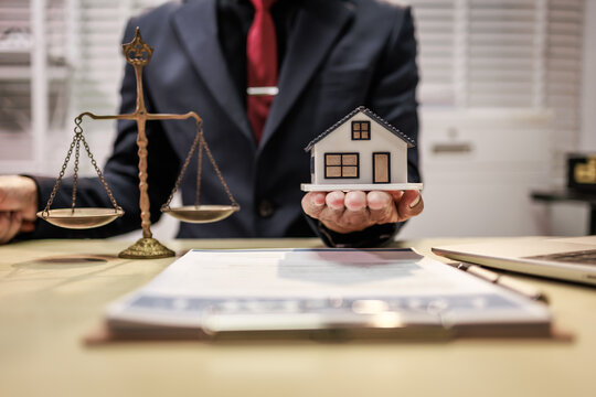 Businessman holding a house model beside scales of justice at a desk, representing real estate law, property rights, and home buying decisions