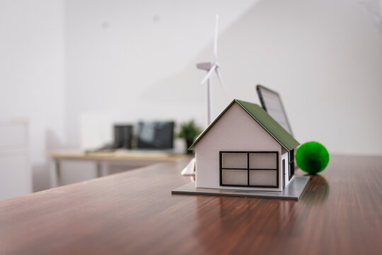 Miniature house model with wind turbine on a wooden desk in a modern office, suggesting eco friendly housing and clean energy planning