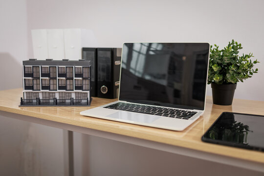 Modern office desk with laptop, tablet, plant, folders and a building model on a wooden table