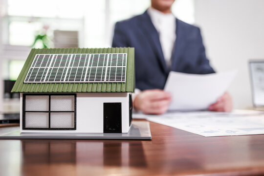 Model house with solar panels on a desk, with a blurred businessman reviewing documents in the background, suggesting green energy and home investment.