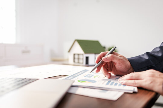 Hands of a real estate agent reviewing charts and plans with a pen beside a small house model and laptop on a desk.