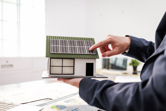 Business person pointing at a small house model with solar panels, green energy, home planning, and sustainable building ideas.