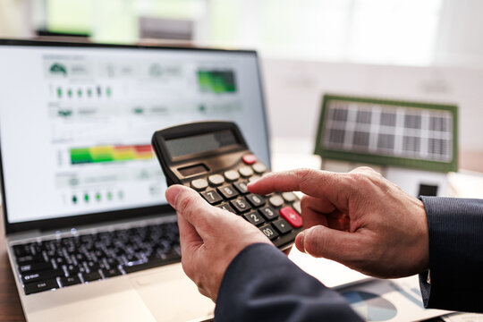Hands using a calculator beside a laptop with charts on screen, suggesting business planning, finance, and solar energy cost analysis in an office setting.