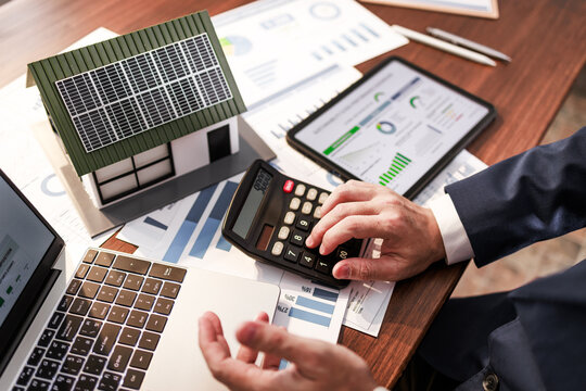 Hands of a businessman using a calculator beside charts, laptop, tablet and a model house with solar panels on a desk.