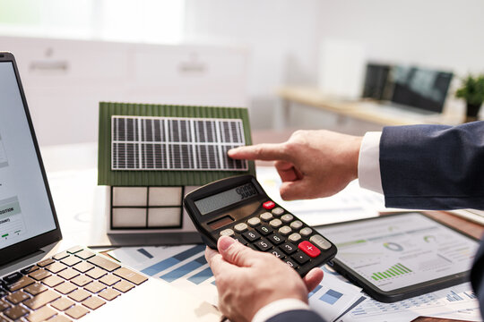 Business person using calculator and pointing at a solar panel model with charts and laptop on a desk, energy planning and investment analysis