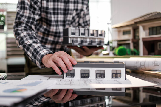 Person holding a house model over architectural plans on a desk, with a modern building in the background