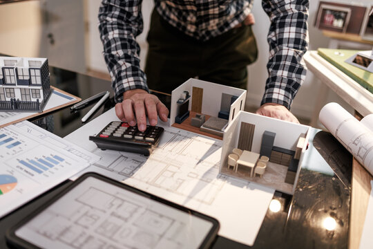Man works on home design plans with calculator, tablet, and miniature room models, interior planning, budgeting, and renovation concept.