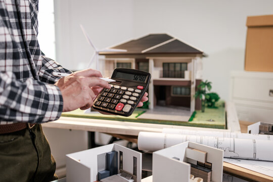Man using a calculator beside a house model and floor plans, real estate budgeting, property planning, and home construction concept.
