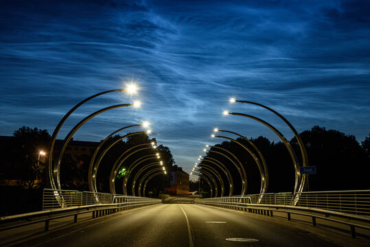 Valmiera Latvia-July 16,2026-Modern illuminated bridge at blue hour with glowing street lights, dramatic clouds, and an empty road creating a striking urban perspective.