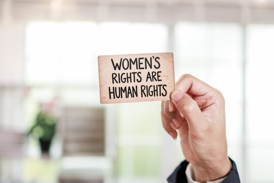 Hand holding a small protest sign with women's rights are human rights text against a blurred indoor background