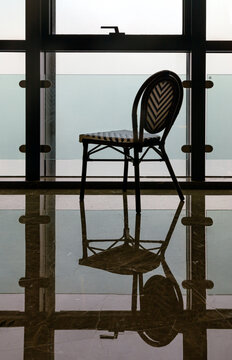 Set against a sweeping, floor-to-ceiling modern glass window, a single woven bistro chair stands in poised tranquility. The chic Parisian-style chair adorned with a classic black-and-cream chevron wea