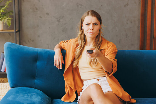 Young annoyed bored woman she 20s wear brown shirt casual clothes watch tv hold remote controller switch channels sit on blue sofa indoor rest at home in own room apartment. People lifestyle concept.