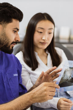 A male dentist in a blue uniform points to a dental x-ray while explaining it to a female patient in a white shirt