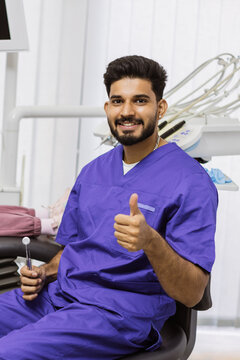 A smiling male dentist in purple scrubs gives a thumbs up while holding a dental mirror in a modern clinic