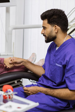 A male dentist in blue scrubs gently shakes a child's hand while holding a dental mirror, reassuring them before a procedure