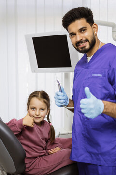 A smiling dentist in blue scrubs gives a thumbs up next to a young girl in a dental chair who also gives a thumbs up