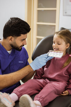 A dentist in blue scrubs and gloves holds a tooth shade guide for a young girl sitting in a dental chair