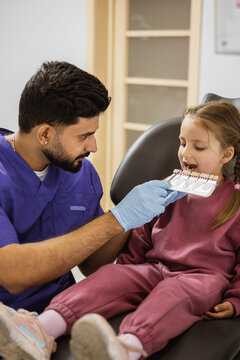 A male dentist in blue scrubs and gloves holds a tooth shade guide up to a young girl's open mouth during a dental examination