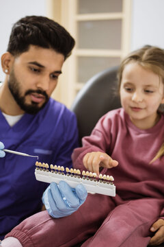 A male dentist in blue scrubs and gloves holds a tooth shade guide for a young girl sitting in a dental chair