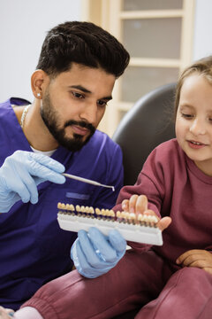 A male dentist in blue scrubs and gloves shows a young girl a shade guide for teeth colors during a dental examination