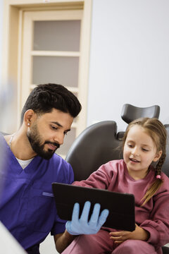 A male dentist in blue scrubs and gloves shows a tablet to a young girl sitting in a dental chair, creating a friendly atmosphere