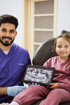 A friendly dentist in blue scrubs and gloves shows a child a dental x-ray on a tablet screen in a modern dental office