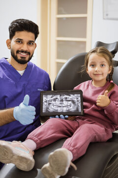 A smiling dentist in scrubs and gloves shows a dental X-ray on a tablet to a happy young girl sitting in a dental chair