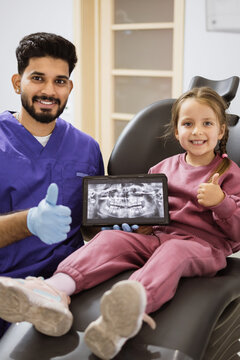 A smiling dentist in scrubs and a young girl give thumbs up while looking at a dental x-ray on a tablet in a modern dental office