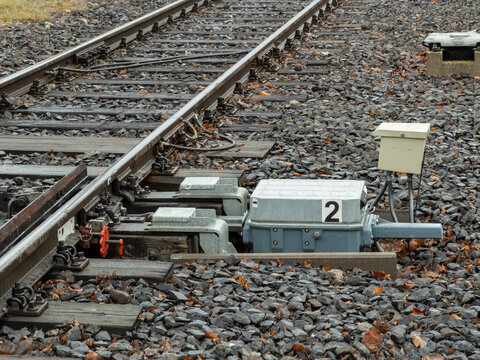 A railway switch point machine mechanism sits beside metal tracks on a gravel bed, ensuring safe train direction changes through automated control systems in a transportation hub