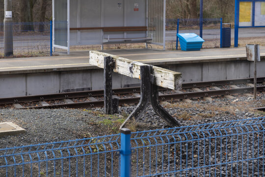 A weathered railway buffer stop stands at the end of a train track, representing industrial safety infrastructure in a quiet rural station setting during a cloudy winter afternoon day