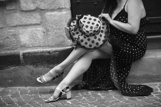Black and white photograph of a woman sitting in a cobblestone street wearing a polka dot dress and holding a matching hat. Elegant urban scene capturing movement, fashion and timeless street photo