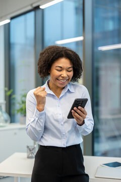 Vertical photo of an African American businesswoman standing outside an office building and rejoicing in success, looking at the mobile phone she is holding in her hand