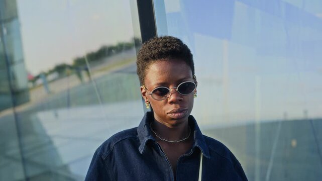Portrait of African American young woman wearing sunglasses, turning head to look at camera near modern glass building. Clip is ideal for diversity marketing and corporate inclusion videos