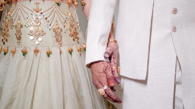 Romantic close up of a South Asian bride and groom holding hands, detailing luxury wedding outfits and henna designs.