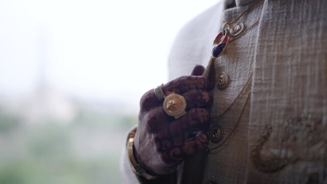 Close up of an Indian groom adjusting his elegant off-white wedding sherwani, showcasing a large gold ring and layered chains.